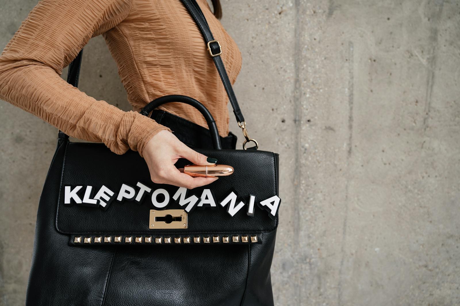 Close-up of a black handbag with 'Kleptomania' letters and a lipstick in hand.